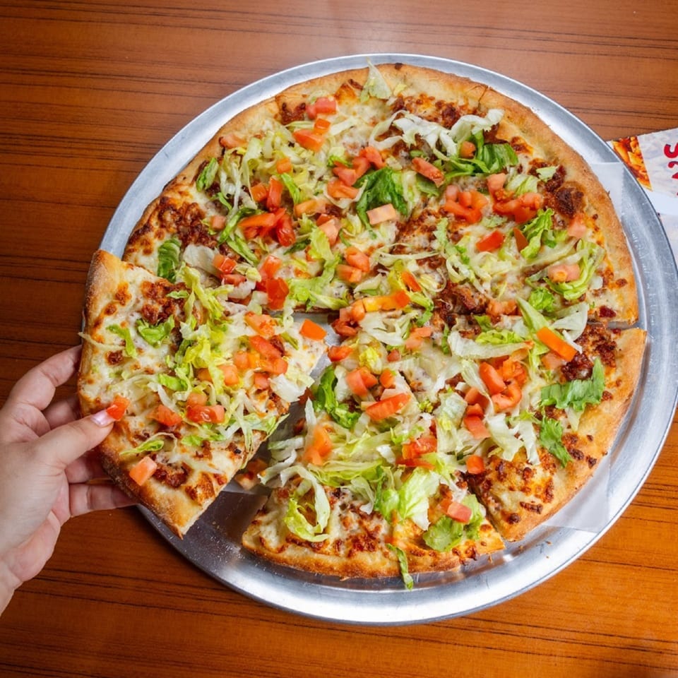 A hand takes a slice of Mama's Pizza & Grill's taco pizza, topped with lettuce and tomatoes.