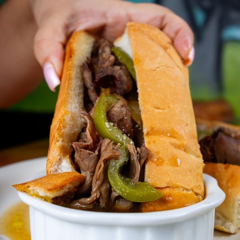 Hand holding a Mama's Pizza & Grill cheesesteak sandwich with green peppers, served with au jus.