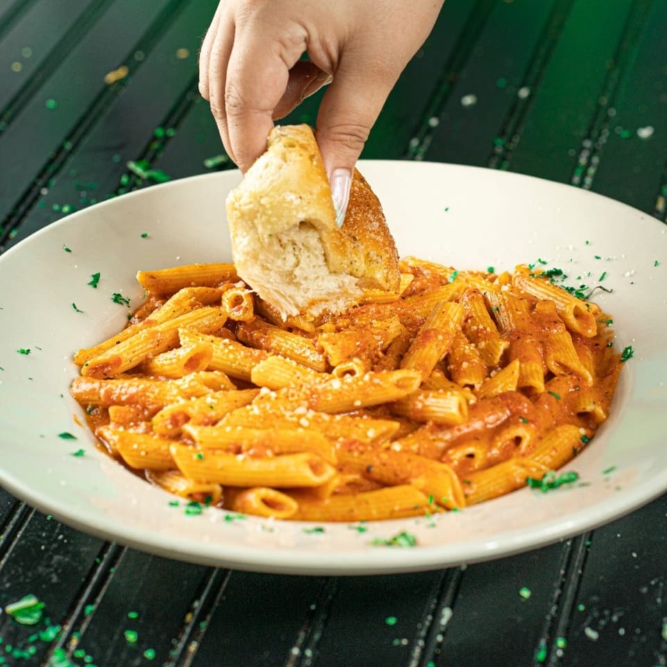 A hand dips a piece of garlic bread into a white bowl of penne pasta with red sauce.